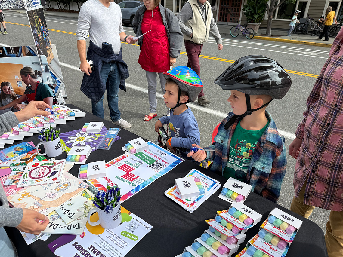 Two children in bike helmets select giveaways at the Anacortes Schools Foundation table at Open Streets 2026. Two children in bike helmets select giveaways at the Anacortes Schools Foundation table at Open Streets 2026.